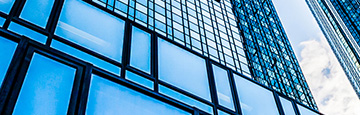 Sliver of an exterior view of windows of two high rises, with a bit of blue sky and clouds visible between and reflected in the windows of the buildings