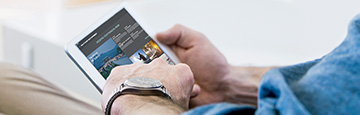 A man’s hand holding a small tablet showing the Voice of the Wealth app home page.
